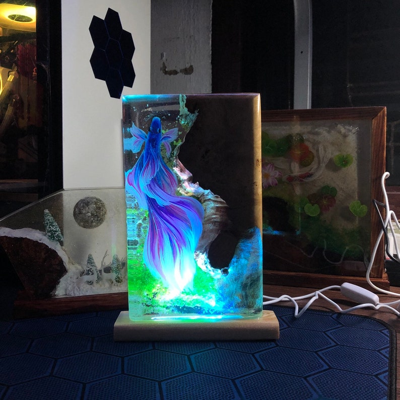 May include: A rectangular resin lamp featuring a vibrant blue and purple betta fish design. The lamp has a wooden base and emits a colorful glow, with the fish appearing to swim in a miniature underwater scene. The lamp is a decorative item.