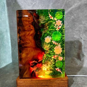 May include: A rectangular lamp with a wooden base and a resin body. The resin is divided into two sections: one with a wood grain pattern and the other with a pond scene featuring koi fish, lily pads, and flowers. The lamp is lit from within.