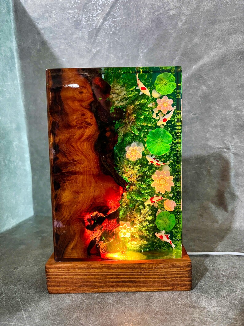 May include: A rectangular lamp with a wooden base and a resin body. The resin is divided into two sections: one with a wood grain pattern and the other with a pond scene featuring koi fish, lily pads, and flowers. The lamp is lit from within.