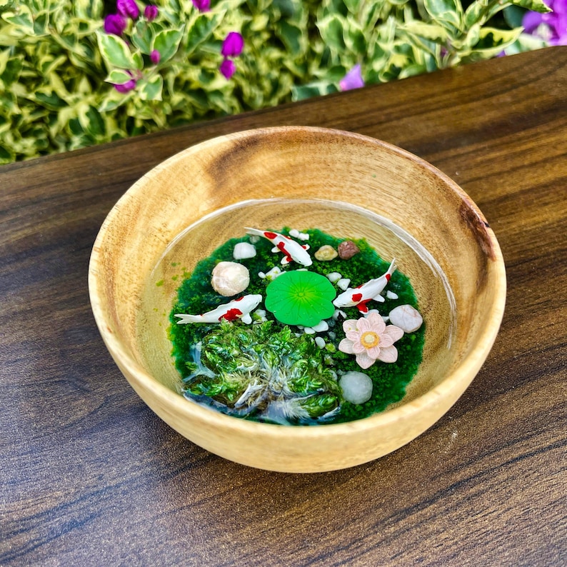 May include: A round wooden bowl containing a clear resin pour that mimics a pond. Inside the resin are miniature white and red koi fish, a bright green lily pad, a pale pink lotus flower, small white pebbles, and patches of green moss.