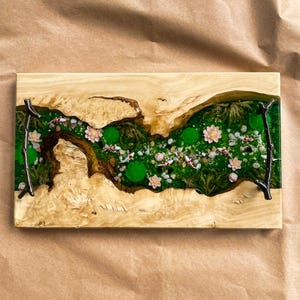 Op de afbeelding: Een rechthoekig houten dienblad met een rivierachtig ontwerp. Het middenstuk heeft groene epoxyhars met miniatuur koikarpers, waterlelies en bloemen. De lade heeft een natuurlijke houtnerf en donkere metalen handgrepen.