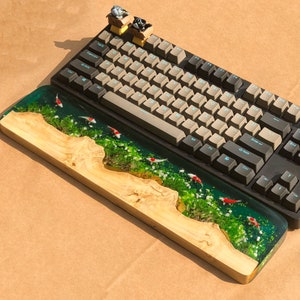 May include: A wooden keyboard wrist rest with a resin insert featuring a green and white landscape with red fish. The wrist rest is sitting on top of a black mechanical keyboard with white keycaps.