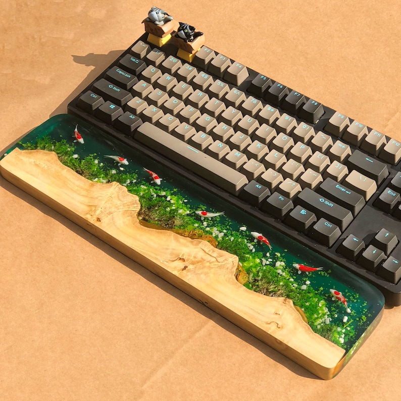 May include: A wooden keyboard wrist rest with a resin insert featuring a green and white landscape with red fish. The wrist rest is sitting on top of a black mechanical keyboard with white keycaps.