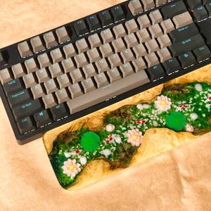 Pu&ograve; includere: Un poggiapolsi in legno per tastiera con un'incisione in resina che rappresenta un fiume verde e bianco con fiori rosa e pesci rossi.
