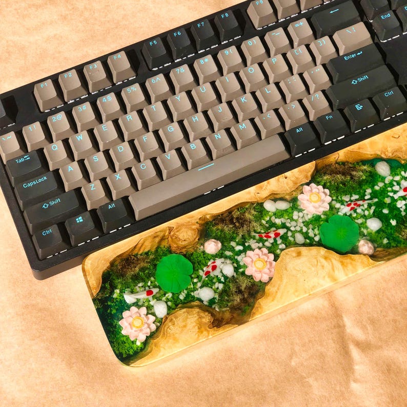 Pu&ograve; includere: Un poggiapolsi in legno per tastiera con un'incisione in resina che rappresenta un fiume verde e bianco con fiori rosa e pesci rossi.