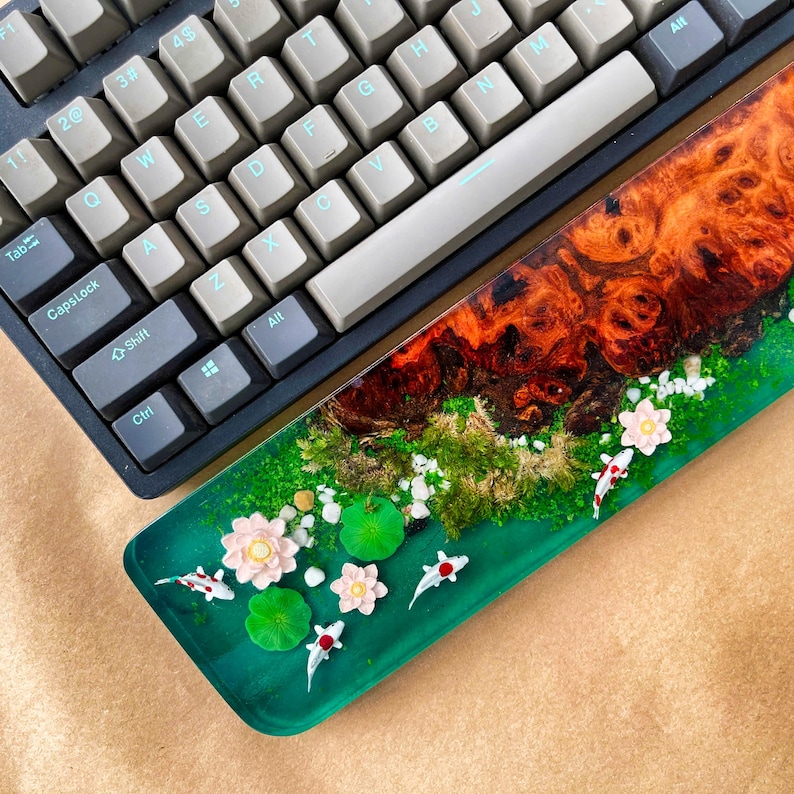 May include: A custom keyboard wrist rest with a unique design. The rest features a vibrant green base with a detailed miniature pond scene, complete with koi fish, lily pads, and flowers. The top section has a wood grain design.