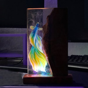 May include: A decorative resin art piece featuring a vibrant betta fish with blue, yellow, and white hues. The fish is encased in clear resin, with a wooden base and a dark, textured side. The piece is illuminated with colorful LED lights.