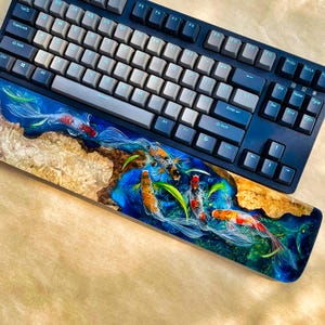 May include: A mechanical keyboard with a custom wrist rest featuring a koi fish design. The keyboard has gray and white keycaps and a dark blue frame. The wrist rest is made of wood and resin, with a vibrant blue and green koi pond scene.