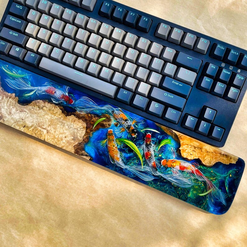 May include: A mechanical keyboard with a custom wrist rest featuring a koi fish design. The keyboard has gray and white keycaps and a dark blue frame. The wrist rest is made of wood and resin, with a vibrant blue and green koi pond scene.