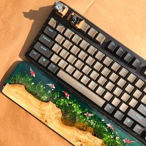 May include: A black mechanical keyboard with a custom keycap set and a wooden wrist rest featuring a resin design with green moss and white fish. The keyboard has a row of function keys labeled F1 through F8.