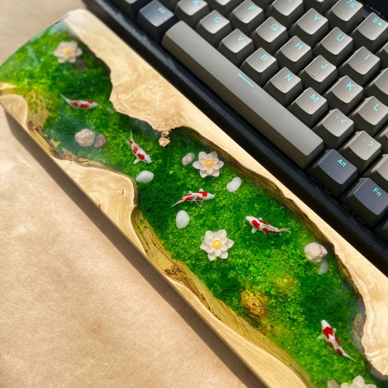 Puede incluir: Un reposamu&ntilde;ecas de teclado de madera con una escena de r&iacute;o. El reposamu&ntilde;ecas presenta un r&iacute;o de resina epoxi verde con peces koi en miniatura, piedras blancas y flores. El teclado es gris con teclas negras.