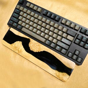 K&ouml;nnte beinhalten: Eine schwarze mechanische Tastatur mit braunen und schwarzen Tastenkappen liegt &uuml;ber einer h&ouml;lzernen Handgelenkauflage. Die Handgelenkauflage hat ein schwarzes Harz-Fluss-Design in hellfarbenem Holz. Die Tastatur hat ein Standard-Layout.