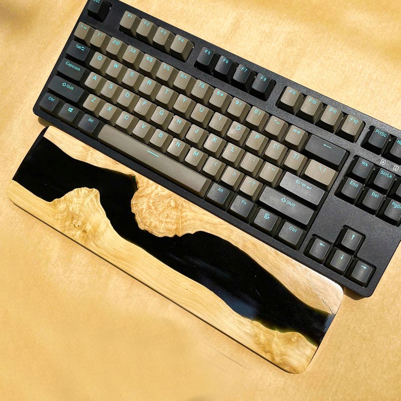 K&ouml;nnte beinhalten: Eine schwarze mechanische Tastatur mit braunen und schwarzen Tastenkappen liegt &uuml;ber einer h&ouml;lzernen Handgelenkauflage. Die Handgelenkauflage hat ein schwarzes Harz-Fluss-Design in hellfarbenem Holz. Die Tastatur hat ein Standard-Layout.