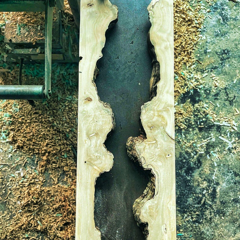 Puede incluir: Una losa de madera clara con bordes naturales y un centro oscuro relleno de resina. La veta de la madera es visible y los bordes tienen una forma natural y org&aacute;nica. Aserr&iacute;n y maquinaria son visibles en el fondo, lo que sugiere un proceso de carpinter&iacute;a.