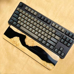 K&ouml;nnte beinhalten: Eine schwarze mechanische Tastatur mit beigefarbenen Tastenkappen und einer h&ouml;lzernen Handgelenkauflage. Die Handgelenkauflage besteht aus hellem Holz mit einem schwarzen Harz-Fluss-Design. Die Tastatur hat ein Standard-Layout mit Funktionstasten.