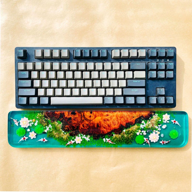 May include: A mechanical keyboard with gray keycaps and a dark blue frame sits above a matching wrist rest. The wrist rest features a vibrant design with a wood grain center, koi fish, white flowers, and green lily pads.
