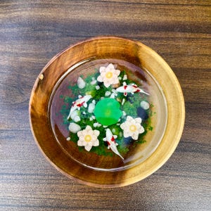 May include: A round wooden bowl containing a clear resin pond scene. Inside the resin are three miniature white and red koi fish, three pale pink lotus flowers, a single bright green lily pad, and scattered small white and grey pebbles.
