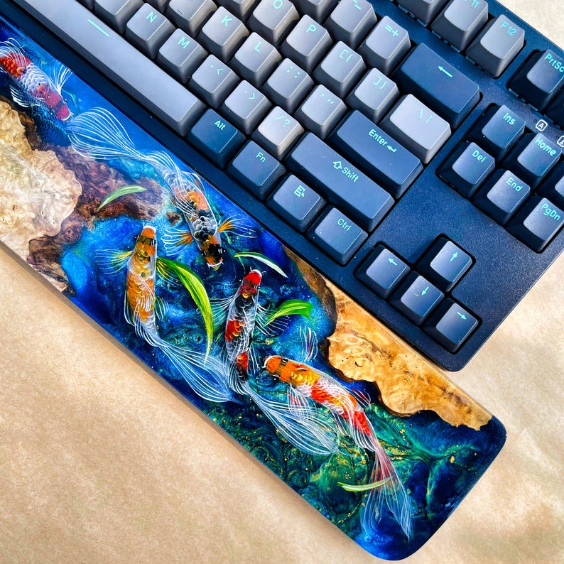 May include: A custom mechanical keyboard with a vibrant koi fish wrist rest. The wrist rest features a detailed resin design with orange, white, and black koi fish swimming in a blue and green water scene. The keyboard has gray and blue keycaps.