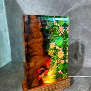 May include: A rectangular lamp with a wooden base. The lamp's body is a combination of wood and resin, featuring a koi pond scene with fish, lily pads, and flowers. The wood has a rich, brown grain. The lamp is lit from within.