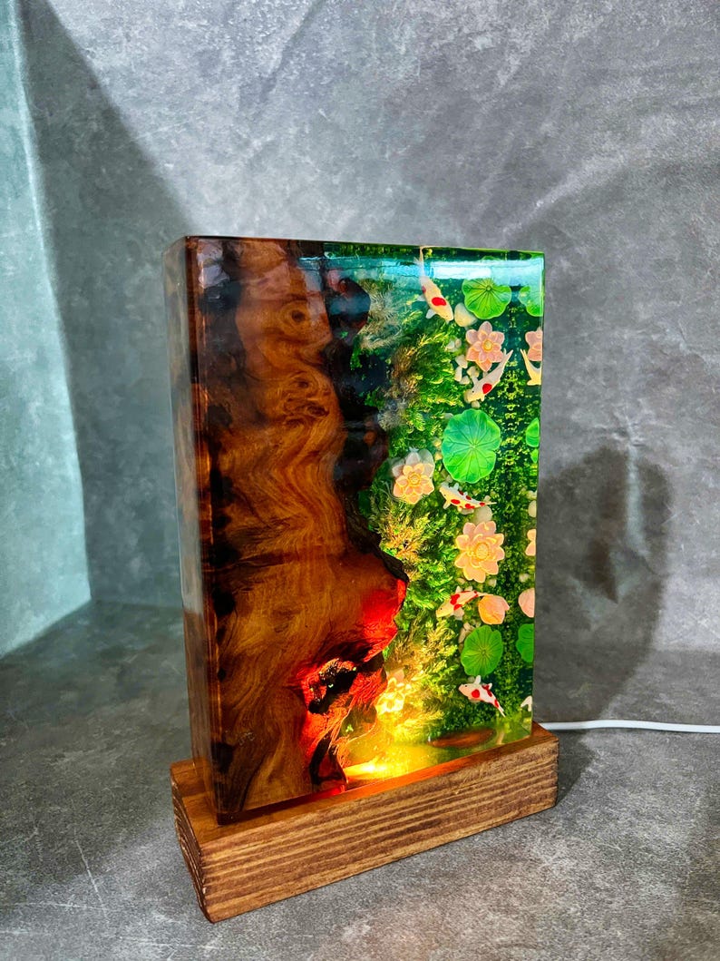 May include: A rectangular lamp with a wooden base. The lamp's body is a combination of wood and resin, featuring a koi pond scene with fish, lily pads, and flowers. The wood has a rich, brown grain. The lamp is lit from within.