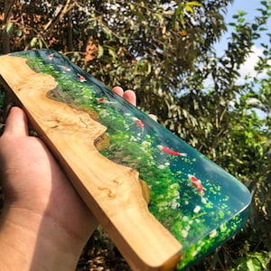 May include: A wooden plaque with a clear resin inlay featuring a green and blue water scene with white flowers and red and white fish swimming.
