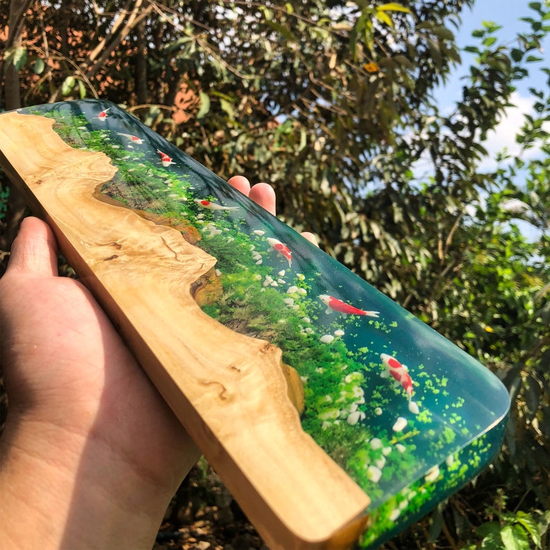 May include: A wooden plaque with a clear resin inlay featuring a green and blue water scene with white flowers and red and white fish swimming.