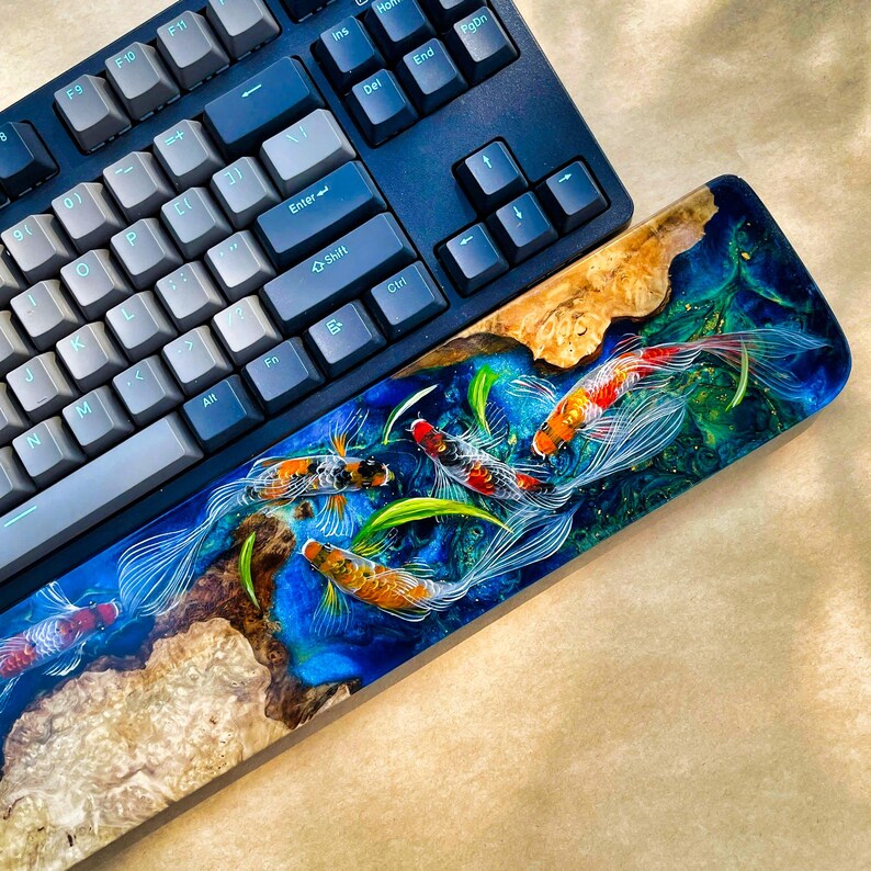 May include: A custom keyboard with a wrist rest featuring a koi fish design. The wrist rest has a blue and green epoxy resin with detailed koi fish illustrations. The keyboard is dark blue with gray and blue keycaps.