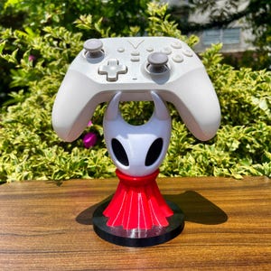 Op de afbeelding: Een witte draadloze videogamecontroller rust op een wit en rood personagevormige standaard. De standaard heeft een wit hoofd met zwarte oogkassen en een rode geplooide basis, gemonteerd op een rond zwart platform voor stabiliteit.