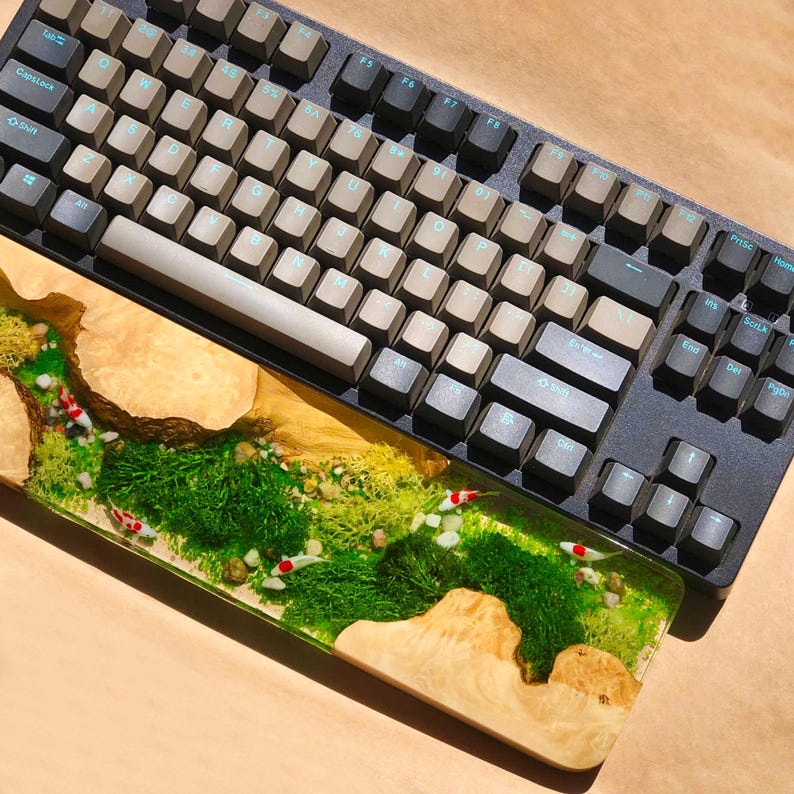 Puede incluir: Un teclado personalizado con un juego de teclas de color marr&oacute;n claro y gris, apoyado en una alfombrilla de escritorio de madera y resina con un dise&ntilde;o de musgo verde y peces blancos.