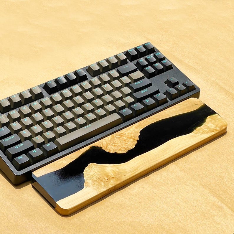 K&ouml;nnte beinhalten: Eine schwarze mechanische Tastatur mit grauen Tastenkappen liegt &uuml;ber einer h&ouml;lzernen Handgelenkauflage. Die Handgelenkauflage hat ein schwarzes Epoxidharz-Flussdesign in hellfarbenem Holz, was eine kontrastreiche &Auml;sthetik erzeugt. Die Tastatur hat ein Full-Size-Layout.