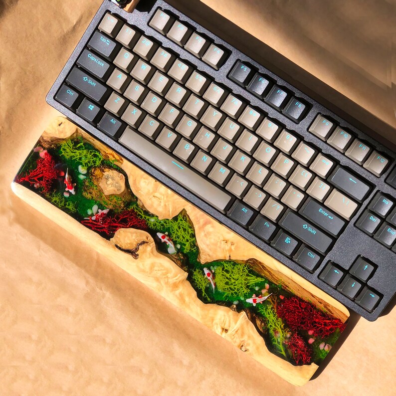 May include: A wooden keyboard wrist rest with a colorful epoxy resin design featuring green, red, and white swirls. The resin design includes small white fish shapes. The wrist rest is placed on top of a black mechanical keyboard with white keycaps.