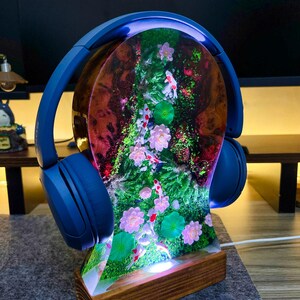 May include: A blue headphone stand with a resin centerpiece featuring a koi pond scene with pink flowers and green lily pads. The stand has a wooden base and a blue arch that holds the headphones.
