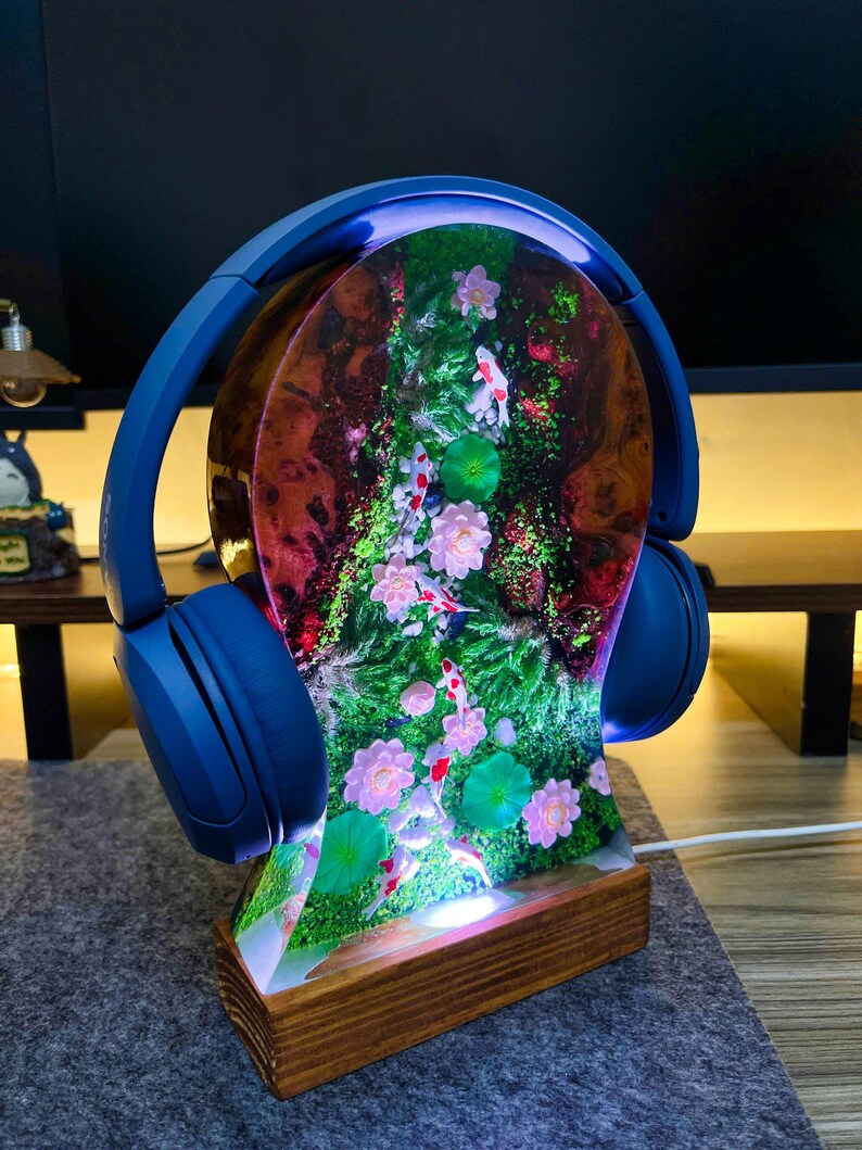 May include: A blue headphone stand with a resin centerpiece featuring a koi pond scene with pink flowers and green lily pads. The stand has a wooden base and a blue arch that holds the headphones.