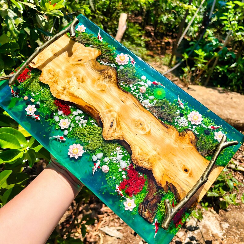 May include: A rectangular serving tray with a unique design. The tray features a wooden centerpiece surrounded by a clear, turquoise resin. The resin encases a miniature garden with koi fish, flowers, and greenery. The tray has bronze-colored handles.