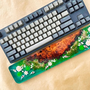 May include: A mechanical keyboard with gray and black keys sits above a custom resin wrist rest. The wrist rest features a wood grain design with a green border, white flowers, and koi fish accents. The keyboard is a dark gray color.