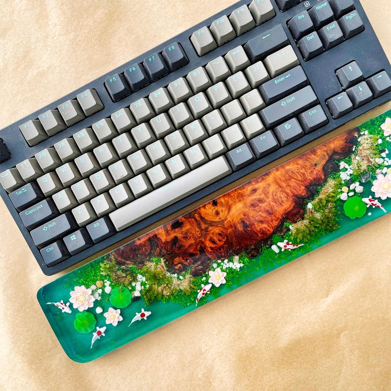 May include: A mechanical keyboard with gray and black keys sits above a custom resin wrist rest. The wrist rest features a wood grain design with a green border, white flowers, and koi fish accents. The keyboard is a dark gray color.
