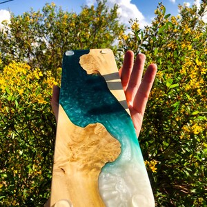 May include: A wooden serving board with a teal and white epoxy resin inlay. The board has a natural wood grain and is finished with a clear coat. The board has four rubber feet on the bottom.
