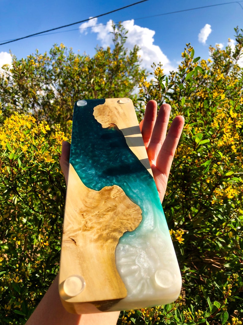 May include: A wooden serving board with a teal and white epoxy resin inlay. The board has a natural wood grain and is finished with a clear coat. The board has four rubber feet on the bottom.