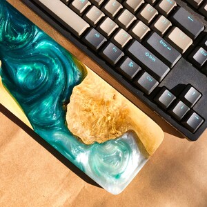 May include: A teal and white resin wrist rest with a wood base. The resin has a swirling pattern and a glittery finish. The wrist rest is sitting on a brown surface next to a black keyboard.
