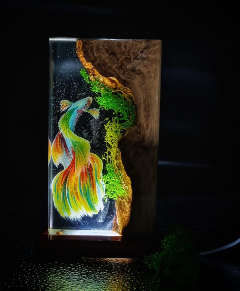 May include: A rectangular resin art piece featuring a colorful betta fish with flowing fins. The fish is set against a dark background with green moss and a wood grain side. The piece is illuminated from below.