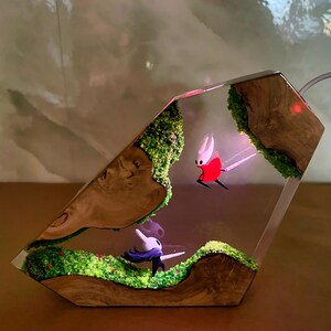 May include: A unique resin art piece featuring two character figurines within a clear, geometric form. The scene includes miniature landscapes with green moss, red berries, and wooden accents. One character is red and white, the other is white and black.