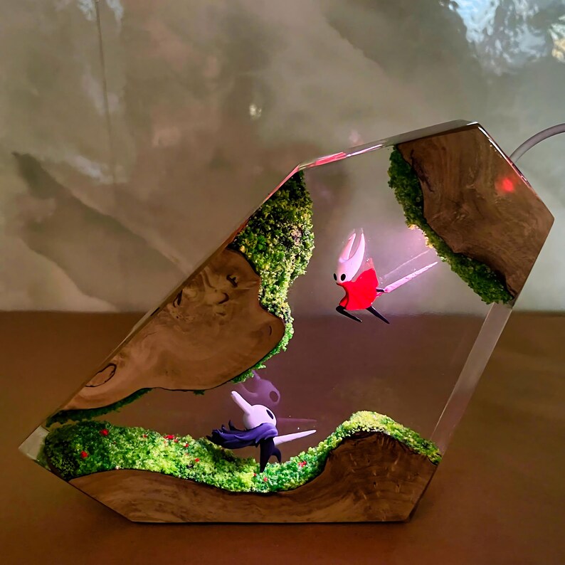 May include: A unique resin art piece featuring two character figurines within a clear, geometric form. The scene includes miniature landscapes with green moss, red berries, and wooden accents. One character is red and white, the other is white and black.