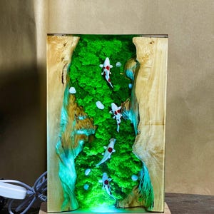 May include: A rectangular wooden lamp with a green moss and resin interior, featuring four koi fish with red and white markings. The lamp has a light-up base and a natural wood grain exterior, creating a unique decorative piece.