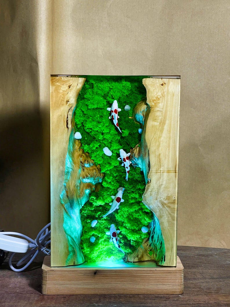May include: A rectangular wooden lamp with a green moss and resin interior, featuring four koi fish with red and white markings. The lamp has a light-up base and a natural wood grain exterior, creating a unique decorative piece.