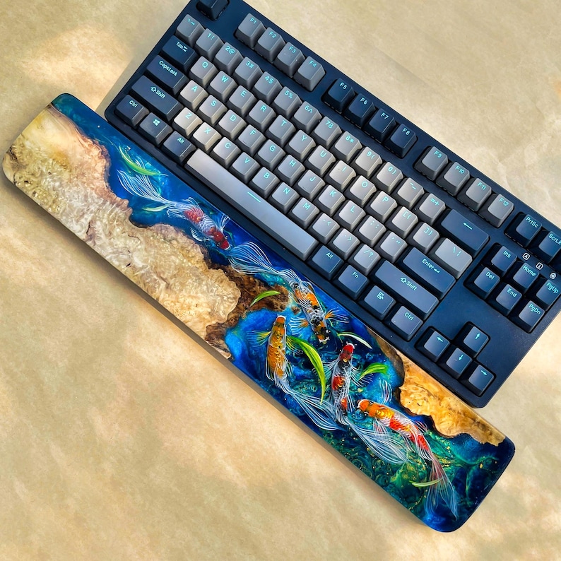 May include: A dark blue mechanical keyboard with gray keycaps rests above a wooden wrist rest. The wrist rest features a koi fish design with blue resin and wood grain.