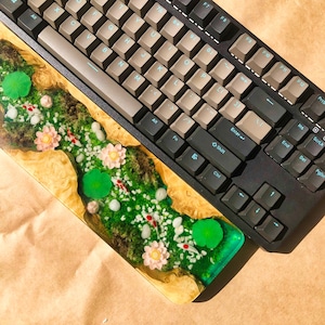 Pu&ograve; includere: Un poggiapolsi in legno per tastiera con un'intarsio in resina che presenta un motivo floreale verde, bianco e rosa. Il poggiapolsi &egrave; posto sopra una tastiera nera.
