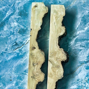 Puede incluir: Dos piezas de madera clara con bordes irregulares y oscuros. La madera tiene un veteado natural y nudos, sobre un fondo azul marmolado. Estas piezas de madera podr&iacute;an usarse para proyectos de artesan&iacute;a o carpinter&iacute;a.