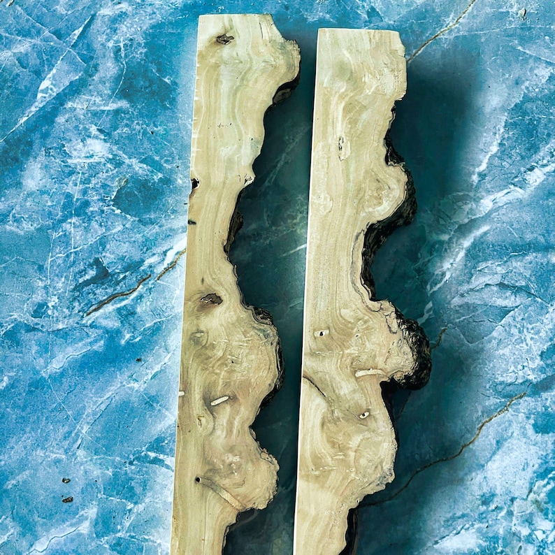 Puede incluir: Dos piezas de madera clara con bordes irregulares y oscuros. La madera tiene un veteado natural y nudos, sobre un fondo azul marmolado. Estas piezas de madera podr&iacute;an usarse para proyectos de artesan&iacute;a o carpinter&iacute;a.
