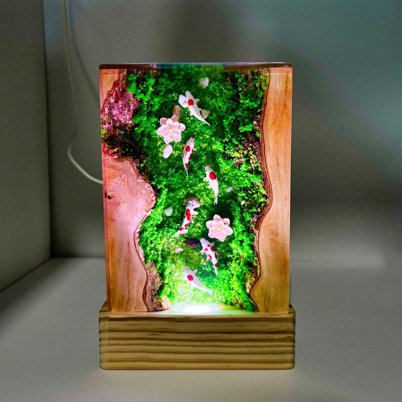Puede incluir: L&aacute;mpara rectangular de madera con un centro de resina transparente. La resina presenta un paisaje de musgo verde con peces koi blancos y rojos y flores rosas. La l&aacute;mpara se asienta sobre una base de madera y emite un brillo suave.