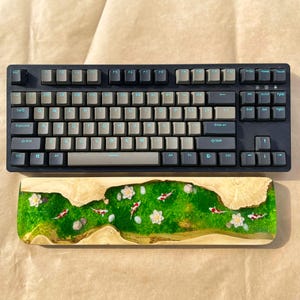 Puede incluir: Un teclado mec&aacute;nico negro con teclas grises y azul claro se encuentra encima de un reposamu&ntilde;ecas de madera. El reposamu&ntilde;ecas presenta un dise&ntilde;o de resina verde y dorada con peces koi, flores blancas y peque&ntilde;as rocas.